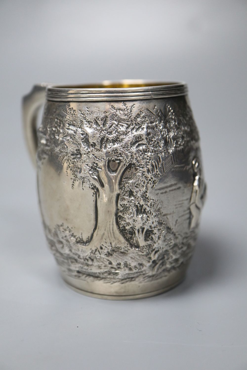 A George III silver barrel shaped christening mug, embossed with figures at play in woodland scene,